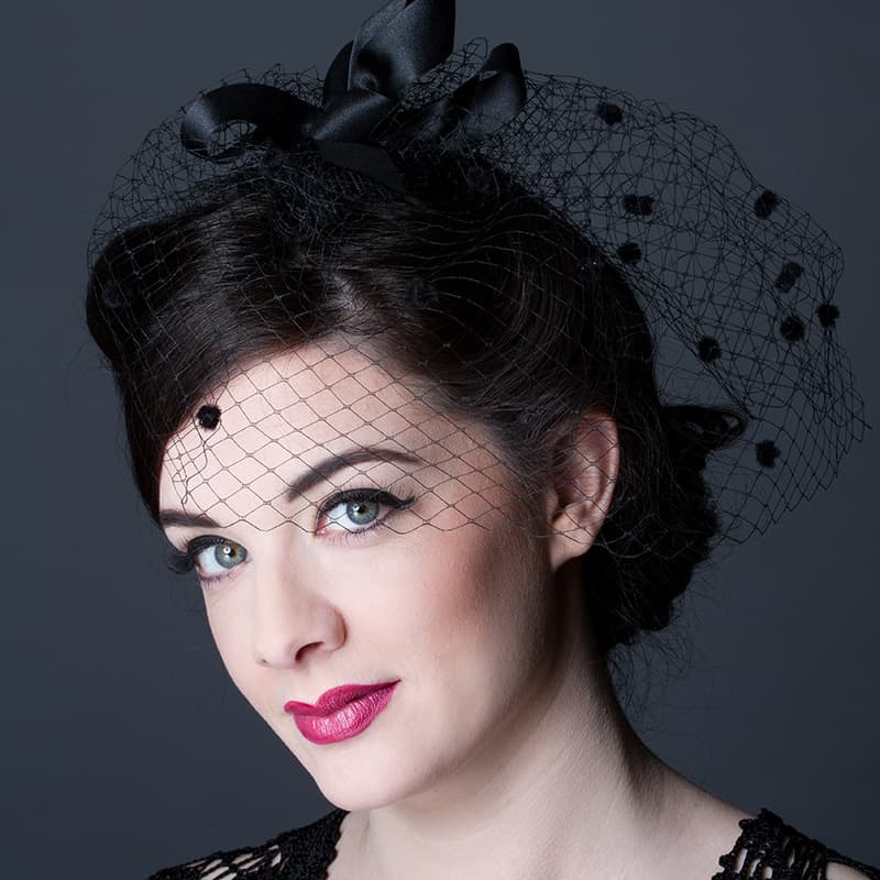 Headshot of woman looking sideways at the camera wearing pink lipstick and a black netted fascinator.