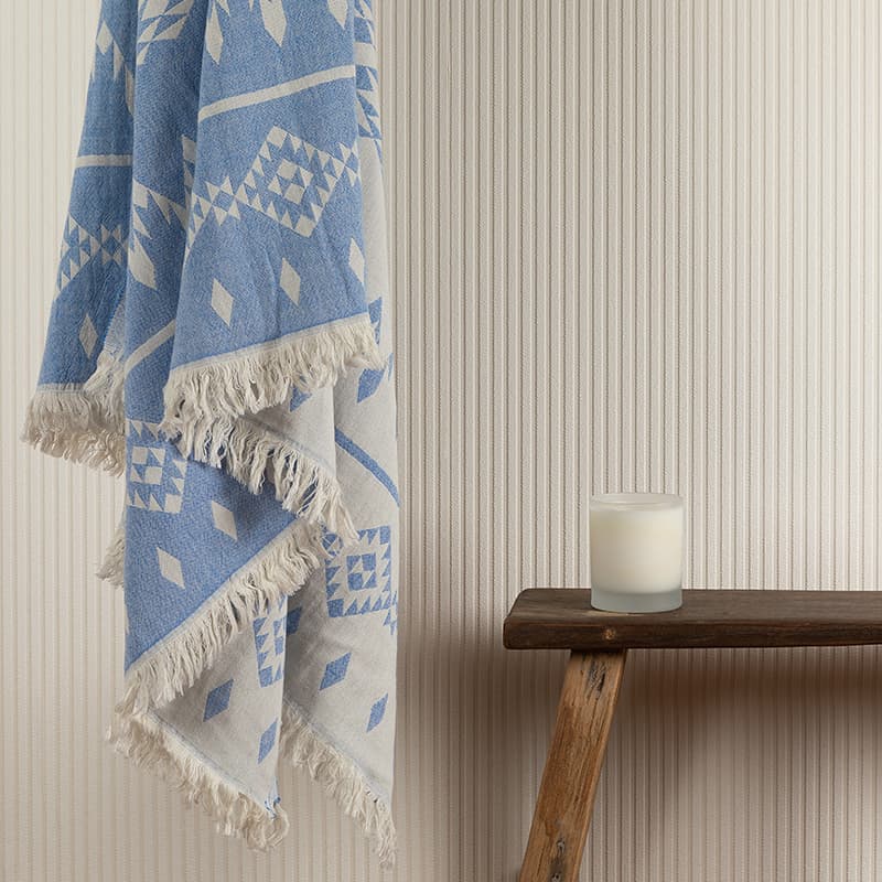 Blue and white throw hanging against a cream pinstripe wall next to a wooden milking stool with a white candle.