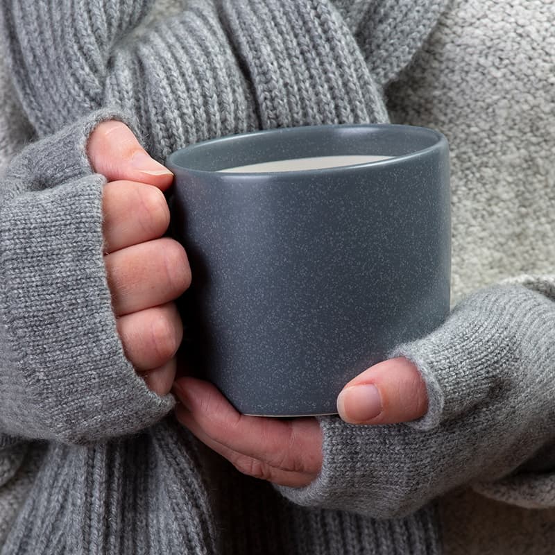 Close up photograph of womens hands in grey fingerless gloves holding a dark grey mug.